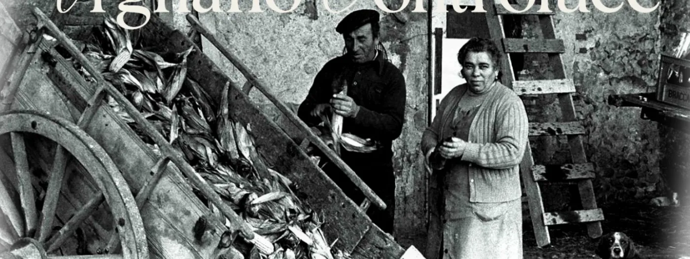Una fotografia in bianco e nero di altri tempi: un uomo con il berretto e una donna davanti a un carro carico di pannocchie, in un cortile di campagna.